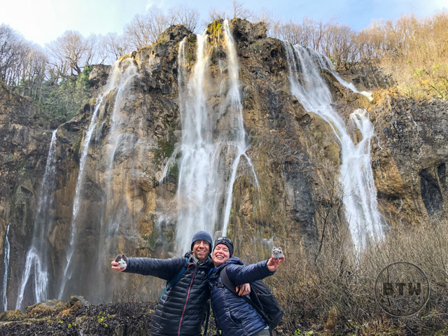 Aaron and Brianna with the travel kitties in front of a large waterfall in Plitvice Lakes National Park, Croatia